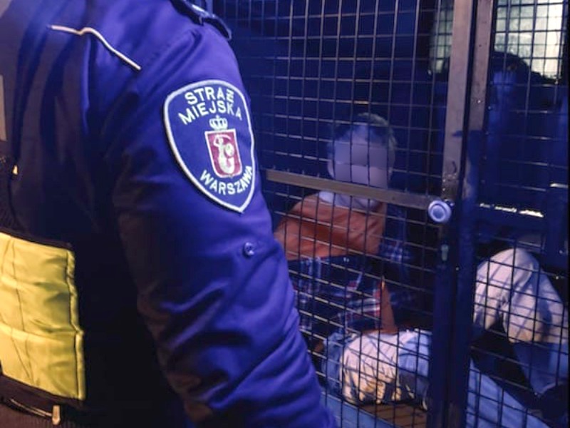 Zdjęcie ilustracyjne: strażnik miejski stojący przy zamkniętym przedziale przewozowym radiowozu. Za kratą siedzi ujęty mężczyzna.