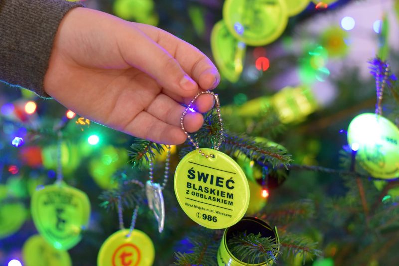 Na zdjęciu widoczna ręka zdejmująca znaczek odblaskowy  z napisem "Świeć blaskiem z odblaskiem" z choinki. W tle gałązki drzewka udekorowane odblaskami.