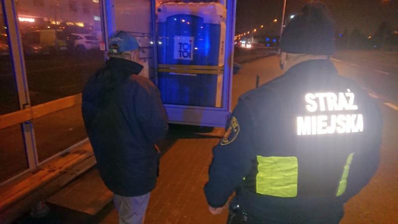 Zdjęcie ilustracyjne: noc, na przystanku stoi zagubiony mężczyzna, obok, w kamizelce z odblaskowym napisem "straż miejska" stoi strażnik.