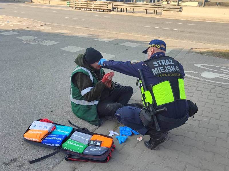 Zdjęcie z interwencji: strażnik miejski kuca przed rannym mężczyzną siedzącym na chodniku, opatrując mu twarz. Obok, na ziemi, leży otwarty plecak ze środkami opatrunkowymi.
