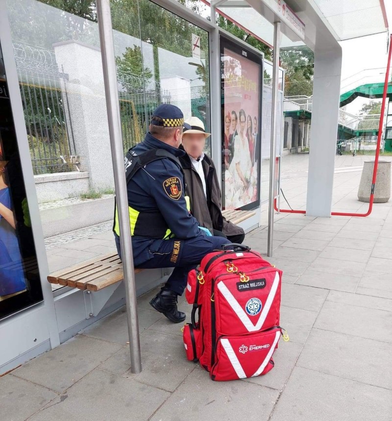 Zagubiony senior siedzący na przystanku. Obok niego strażnik miejski, na ziemi stoi plecak ratowniczy.