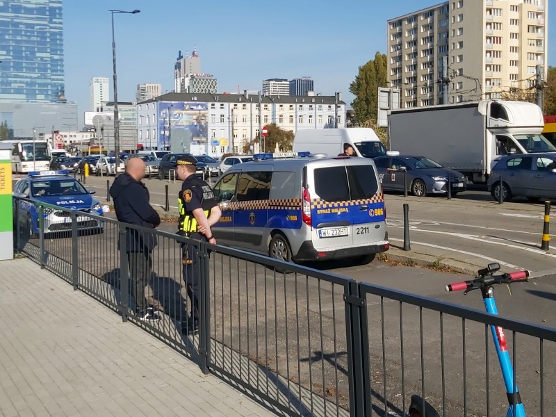 Strażnik miejski stoi obok opartego o barierki mężczyzny w czarnej kurtce. Obok nich stoi radiowóz straży miejskiej, za nimi- samochód policyjny.