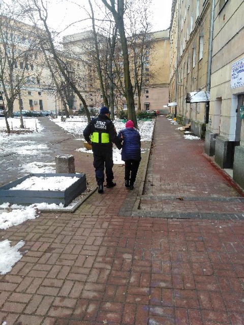 Starsza kobieta prowadzona pod ramię przez strażnika miejskiego.