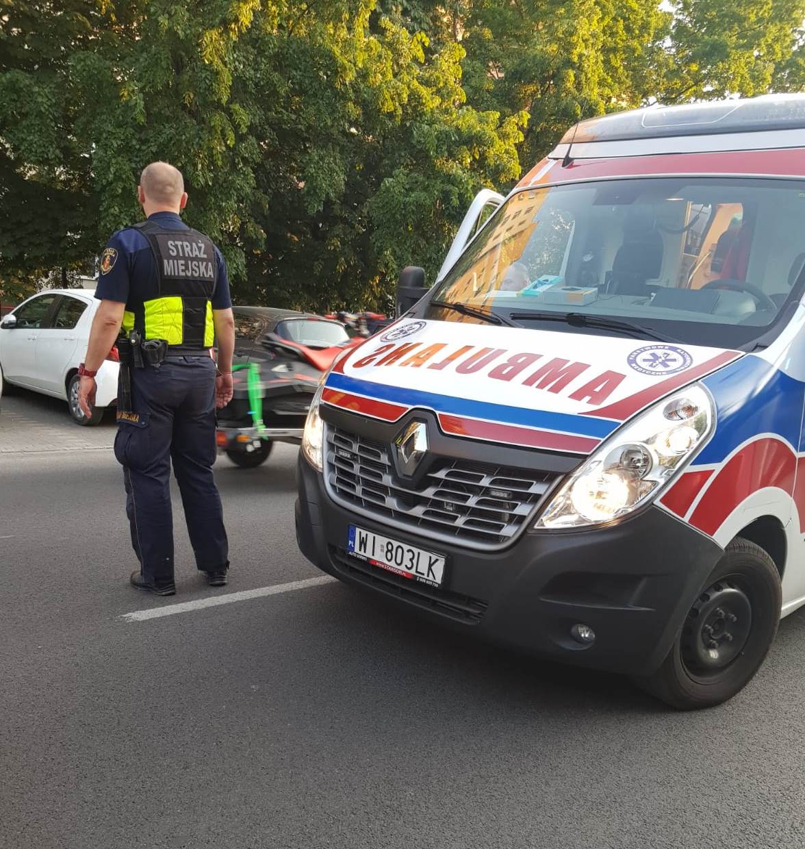 Strażnik miejski, stojąc na jezdni obok ambulansu, zabezpiecza miejsce pracy zespołu ratowniczego.