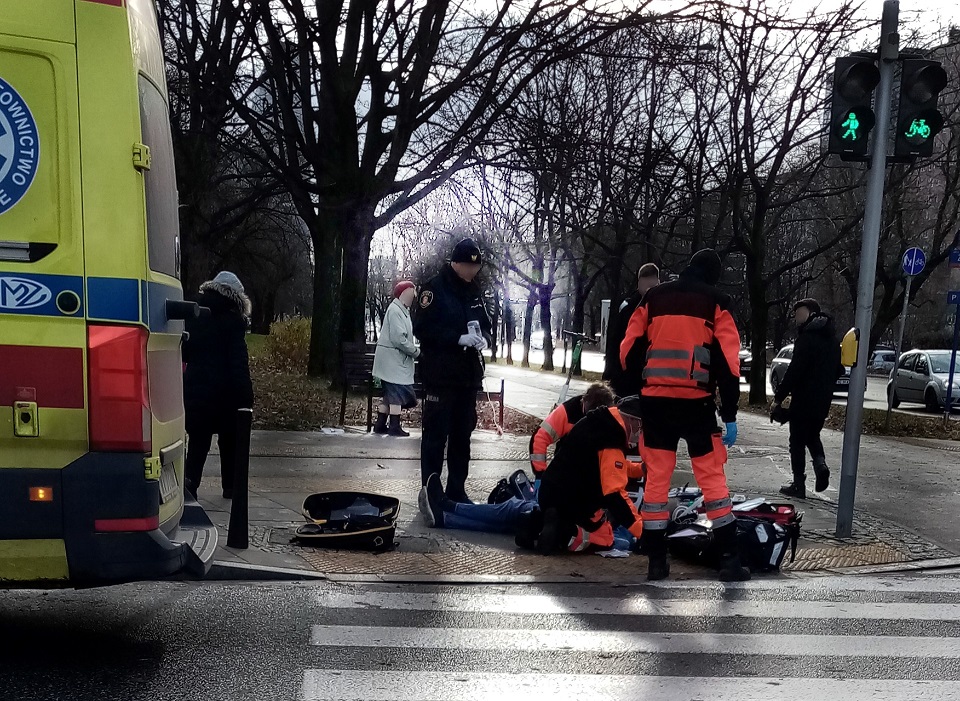 Ratownicy i strażnik miejski prowadzą akcję resuscytacyjną mężczyzny leżącego na skraju chodnika.
