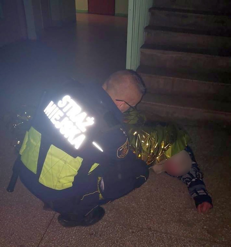 Strażnik pochylający się nad leżącym na klatce schodowej mężczyzną okrytym folią termiczną