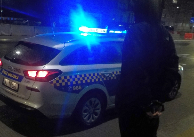 Radiowóz straży miejskiej nocą, obok stoi mężczyzna w ciemnym ubraniu, zakuty w kajdanki.