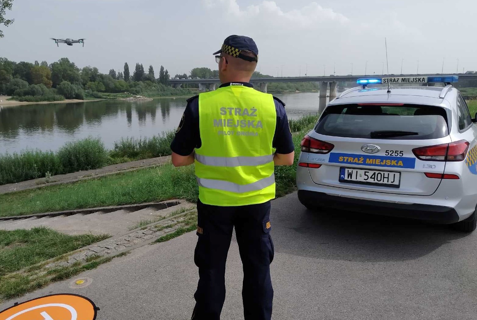 Strażnik miejski w trakcie oblotu dronem okolic jeziorka Gocławskiego. Dron w locie, strażnik z urządzeniem sterującym i radiowóz straży miejskiej nad wodą.