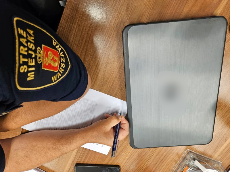 Laptop leżący na biurku, obok strażnik miejski piszący notatkę służbową- zdjęcie ilustracyjne