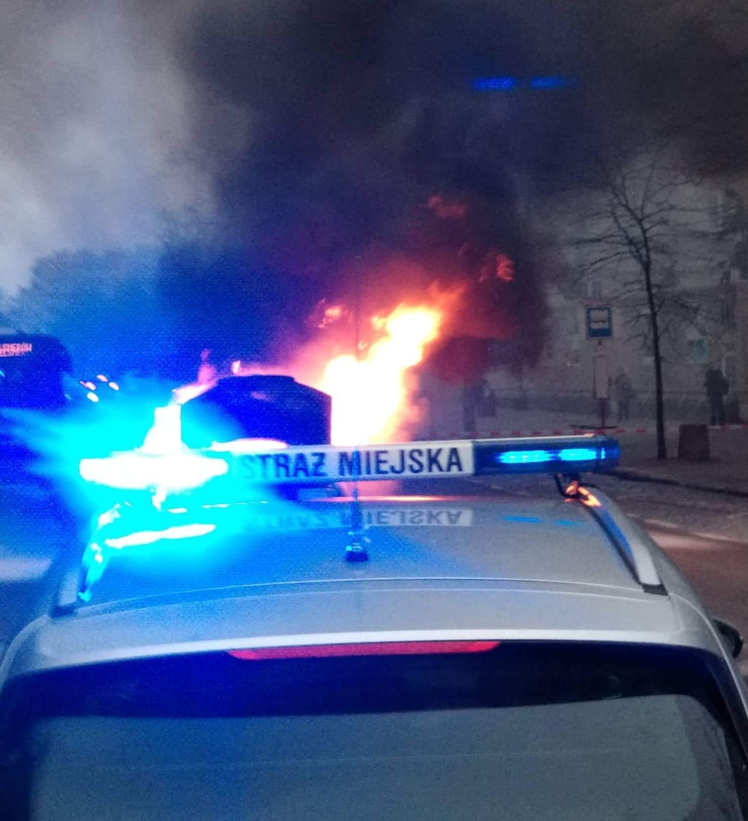 Zdjęcie ilustracyjne: radiowóz straży miejskiej z włączonymi światłąmi sygnalizacyjnymi. Za nim widoczne płomienie i kłęby dymu z płonącego samochodu.