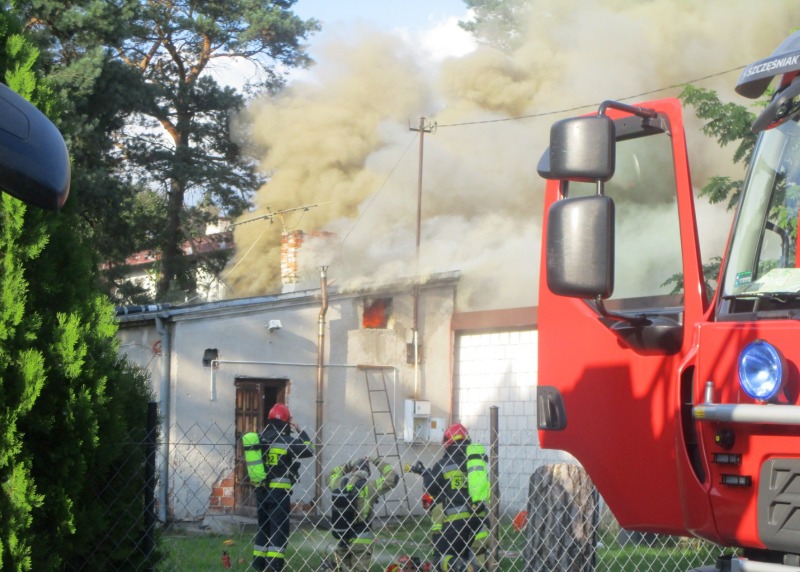 Widok na płonący budynek, z którego unoszą się kłęby dymu. Przed budynkiem trzech strażaków podczas akcji gaśniczej. Na pierwszym planie, z prawej strony kadru, szoferka samochodu strażackieho.