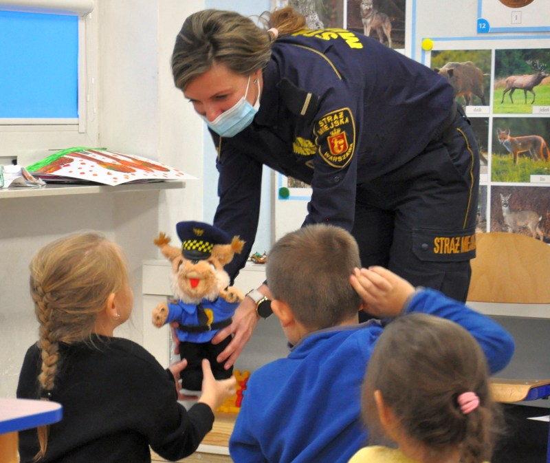 Zdjęcie ilustracyjne: strażniczka miejska podczas zajęć z dziećmi wręcza maskotkę Straży Miejskiej dziewczynce siedzącej w ławce szkolnej.