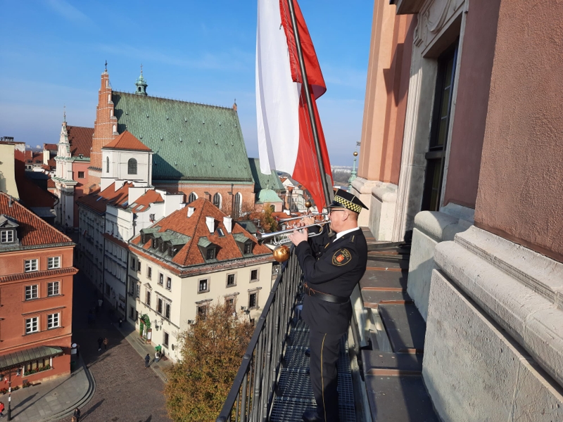 Hejnalista straży miejskiej na wieży Zamku Królewskiego wykonuje hymn państwowy. W tle powiewa biało-czerwona flaga.