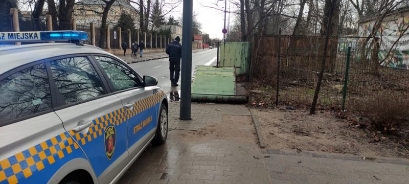 Zdjęcie z interwencji: radiowóz straży miejskiej przy przewróconej bramie. Obok strażnik miejski zabezpieczający miejsce zdarzenia.