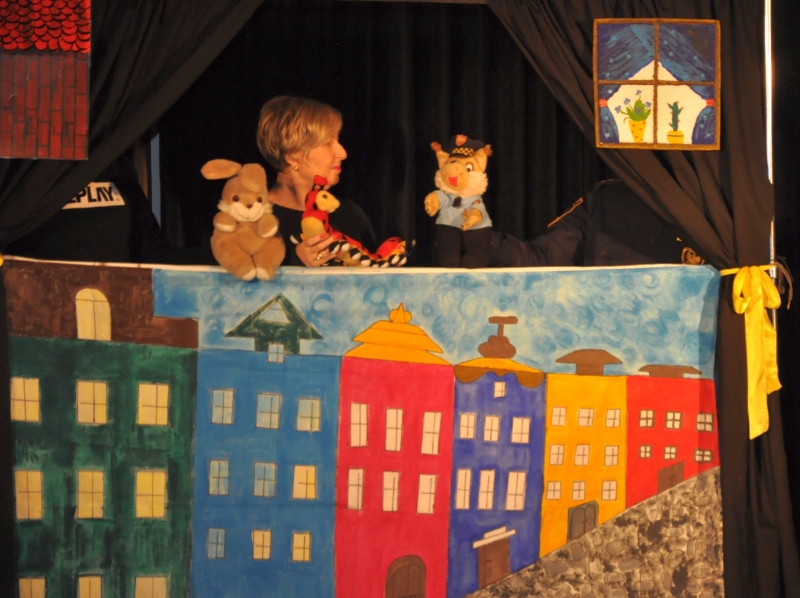Zdjęcie z przedstawienia: zza zasłony przedstawiającej kolorowe, rysunkowe domy stoją strażnicy-aktorzy, trzymający postaci z bajki.