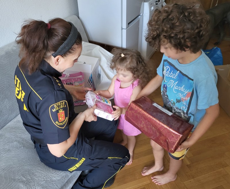 Strażniczka miejska podająca dwie paczki dzieciom z Ukrainy: młodsza dziewczynka w różowej sukience odbiera paczkę z akcesoriami do włosów, starszy chłopiec, w niebieskiej koszulce, trzyma w rękach owiniętą w błyszczący czerwono-brązowy papier paczkę.