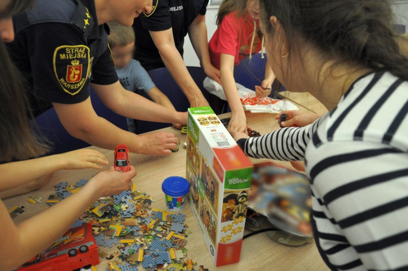 Grupa dzieci bawiąca się puzzlami rozsypanymi na stole. Obok nich strażniczka miejska.