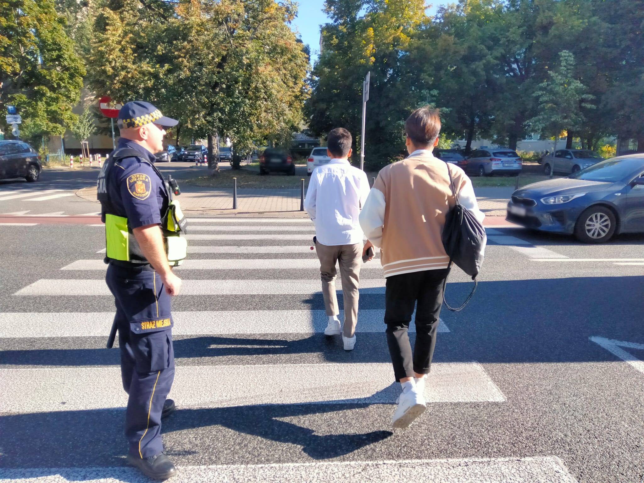 Strażnik miejski na przejściu dla pieszych zabezpiecza spokojne przekroczenie jezdni przez dwóch młodzieńców