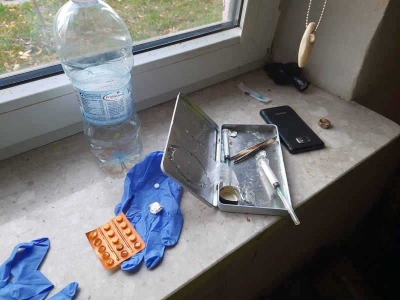 Blaszane pudełko ze strzykawką, listek z tabletkami, niebieskie rękawiczki jednorazowe, telefon i butelka z wodą na parapecie okiennym.