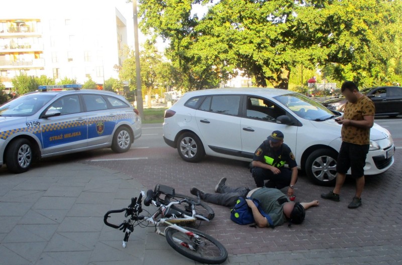 Strażnik miejski pochylający się nad leżącym na ziemi rowerzystą. Obok stoi kierowca białego renault.